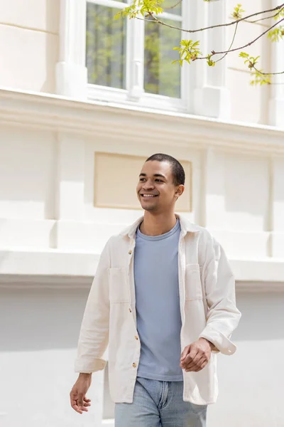 Young and cheerful african american man in shirt jacket walking in urban city — Stock Photo