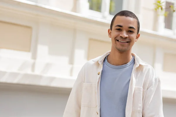 Joven y feliz afroamericano en camisa chaqueta sonriendo en ciudad urbana - foto de stock