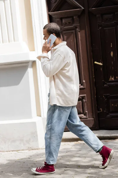 Longitud completa de hombre afroamericano en camisa chaqueta hablando en el teléfono móvil en la calle en la ciudad urbana - foto de stock