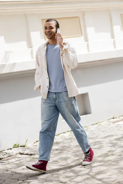 Longitud completa de sonriente hombre afroamericano en camisa chaqueta hablando en el teléfono móvil en la calle en la ciudad urbana - foto de stock