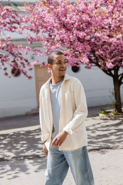 Positiver afrikanisch-amerikanischer Mann in Hemdjacke, der in der Nähe eines rosafarbenen Kirschbaums spaziert — Stockfoto