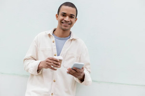 Hombre afroamericano feliz sonriendo mientras sostiene teléfono inteligente y taza de papel afuera - foto de stock