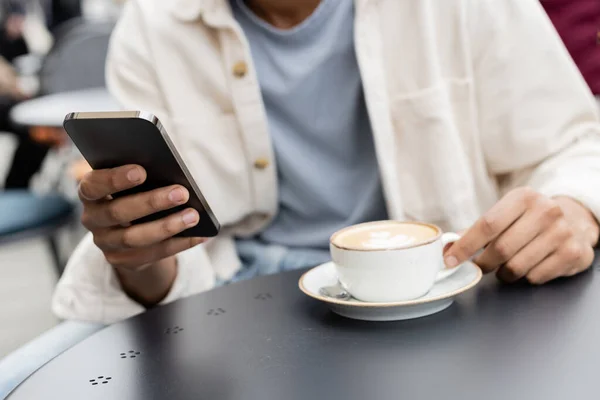 Visão parcial do homem afro-americano mensagens de texto no smartphone perto cappuccino na mesa — Fotografia de Stock