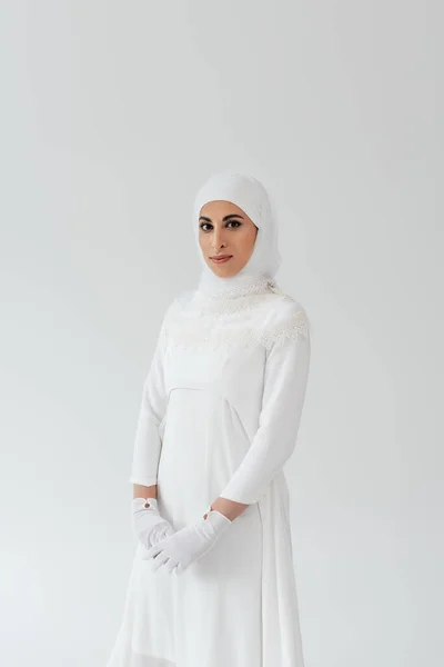 Joven novia musulmana en hijab y vestido blanco mirando a la cámara aislada en gris - foto de stock