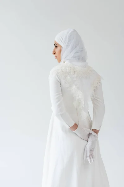 Muslim bride in gloves and white dress standing with hands behind back isolated on grey — Stock Photo