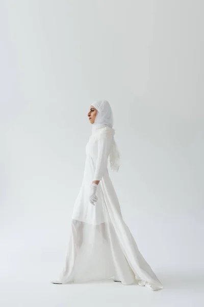 Vista lateral de la novia musulmana en hijab y vestido blanco caminando sobre gris - foto de stock