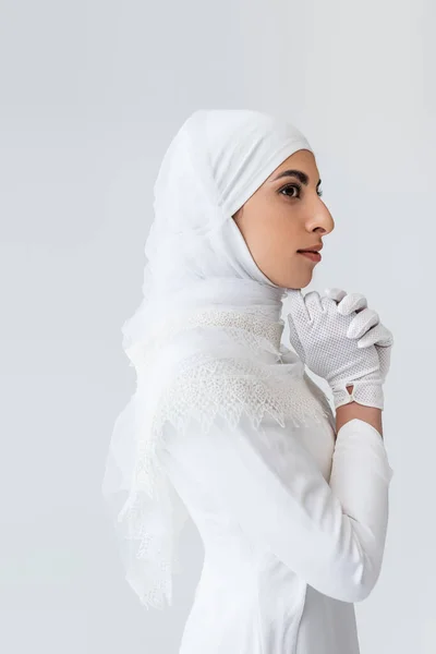 Молодая мусульманская невеста в перчатках и свадебном платье, молящаяся изолированно на серой — стоковое фото