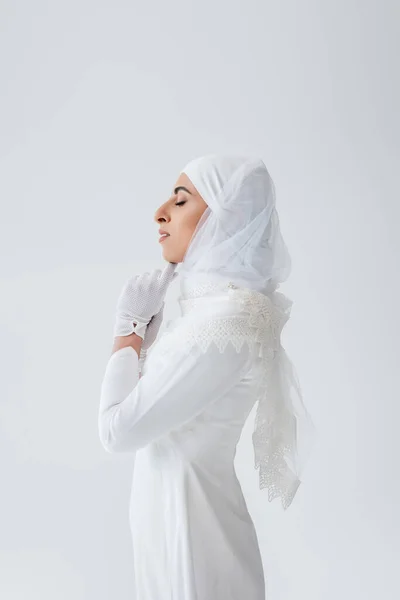 Side view of muslim bride in gloves and wedding dress praying isolated on grey — Stock Photo