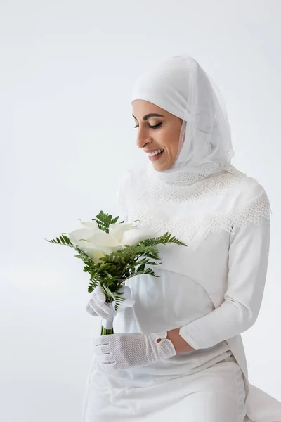 Happy muslim bride in gloves and wedding dress holding calla lily flowers isolated on grey — Stock Photo