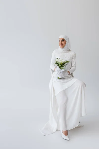 Full length of cheerful muslim bride in gloves and wedding dress holding calla lily flowers and sitting on chair on grey — Stock Photo