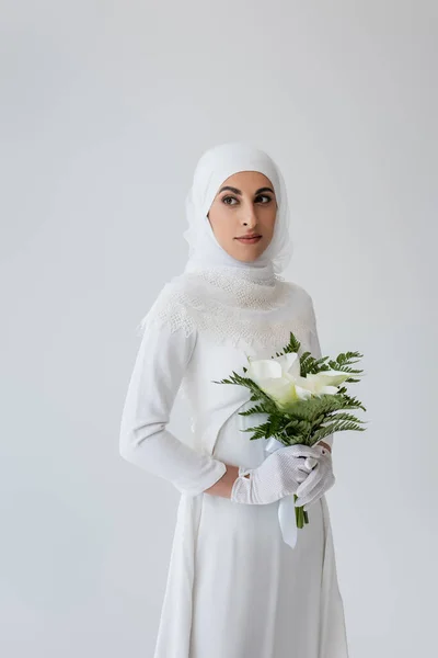Muslim bride in gloves and wedding dress holding bouquet of calla lily isolated on grey — Stock Photo