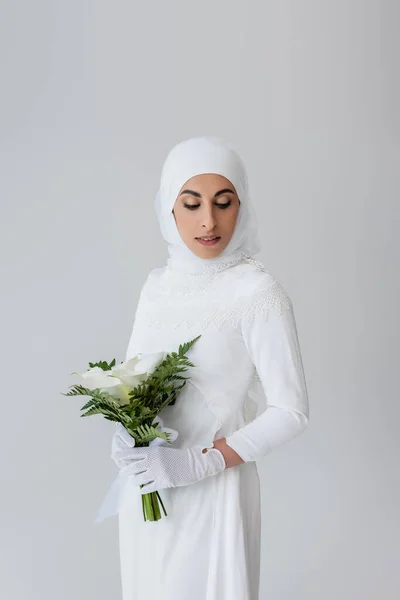 Novia musulmana en guantes y vestido blanco sosteniendo flores de lirio de cala aisladas en gris - foto de stock