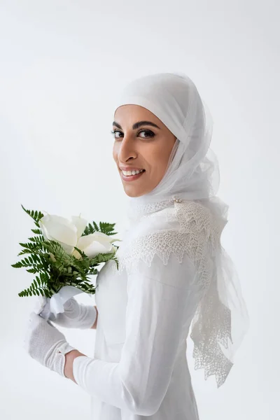 Весёлая мусульманская невеста в перчатках и свадебном платье с цветами калла лилия, изолированными на сером — стоковое фото