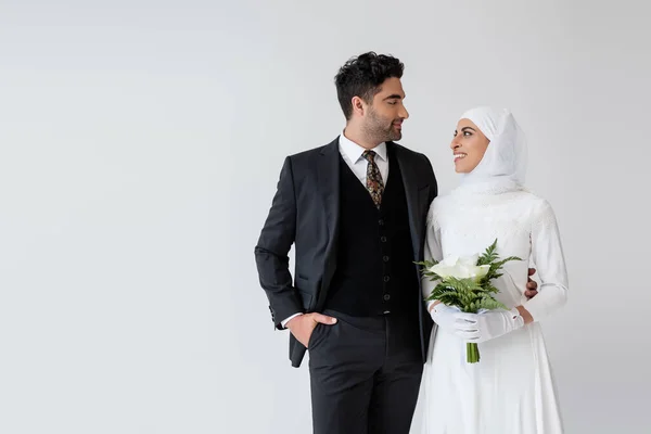 Мусульманская невеста в свадебном платье держит букет калла лилии и смотрит на счастливого жениха в костюме, изолированном на сером — стоковое фото