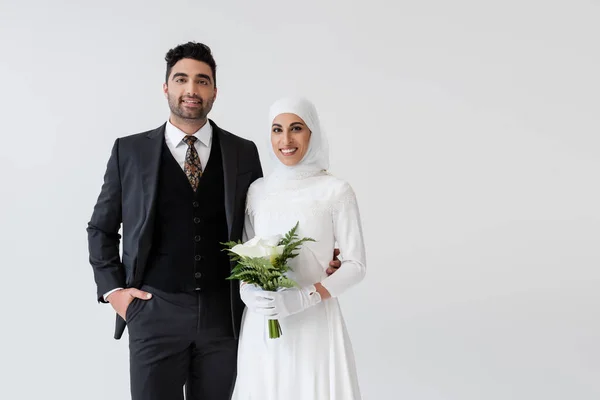 Мусульманская невеста в перчатках и свадебном платье с букетом калла лилии рядом с счастливым женихом в костюме, изолированном на сером — стоковое фото