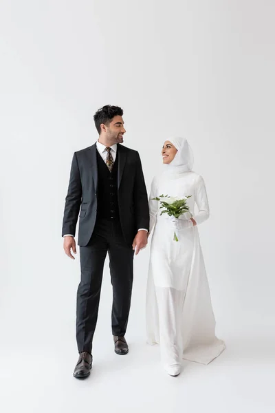 Muslim bride in wedding dress holding bouquet of calla lily and looking at happy groom in suit on grey — Stock Photo