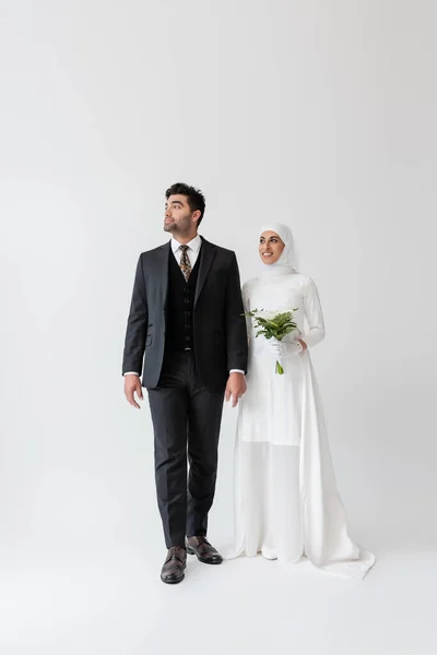 Muslim bride in wedding dress holding bouquet of calla lily and standing with groom in suit on grey — Stock Photo