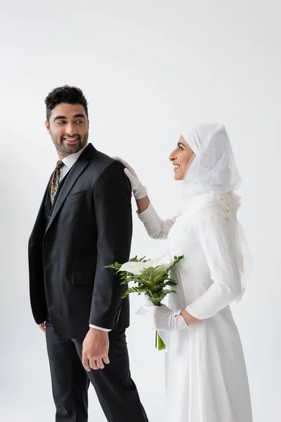 Щаслива мусульманська наречена у весільній сукні тримає букет каллі та стоїть за нареченим у костюмі ізольовано на сірому — стокове фото