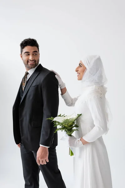 Happy muslim bride in wedding dress holding bouquet of calla lily and standing behind groom in suit isolated on grey — Stock Photo