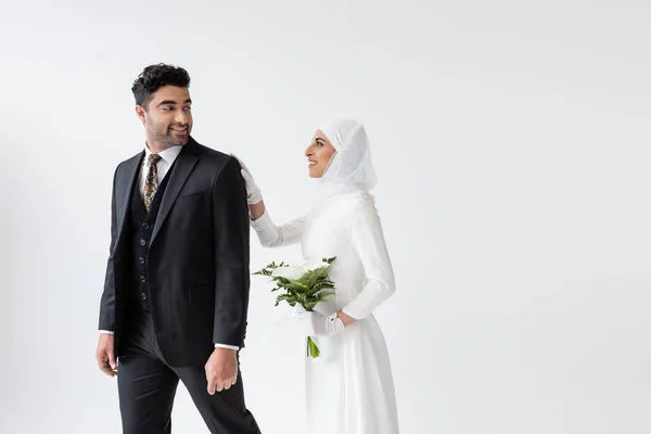 Lächelnde muslimische Braut im Brautkleid mit einem Bouquet Calla-Lilie und hinter dem Bräutigam im Anzug isoliert auf grau — Stockfoto