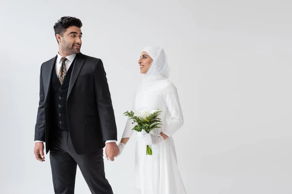 Улыбающаяся мусульманская невеста в свадебном платье с букетом калла лилия, держащаяся за руки с женихом в костюме, изолированном на сером — стоковое фото