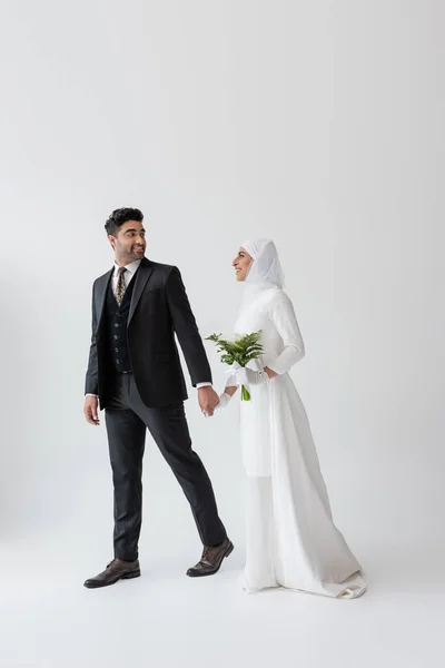 Novia musulmana sonriente en vestido de novia con ramo de lirio de cala cogido de la mano con el novio en traje en gris - foto de stock