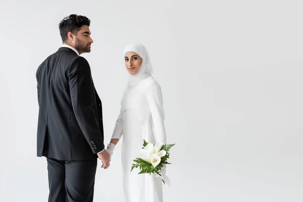 Lächelnde muslimische Braut im Brautkleid mit Bouquet von Calla Lilie Händchen haltend mit Bräutigam isoliert auf grau — Stockfoto
