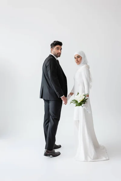 Noiva muçulmana sorridente em vestido branco com buquê de lírio calla de mãos dadas com o noivo em cinza — Fotografia de Stock