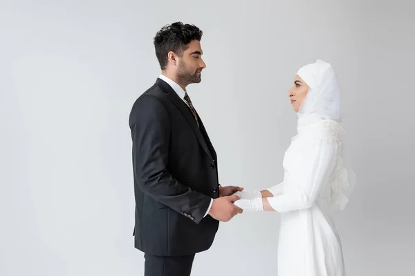 Вид сбоку мусульманского жениха и невесты, держащихся за руки и смотрящих друг на друга, изолированных на сером — стоковое фото