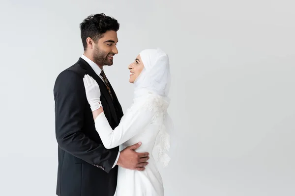 Happy muslim bride in hijab and wedding dress and groom hugging while looking at each other isolated on grey — Stock Photo