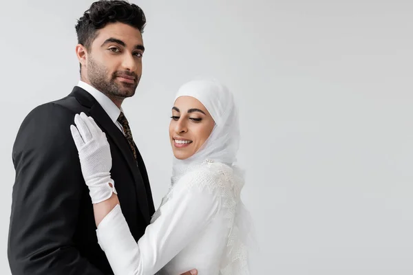 Happy muslim bride in hijab and wedding dress and groom hugging isolated on grey — Stock Photo