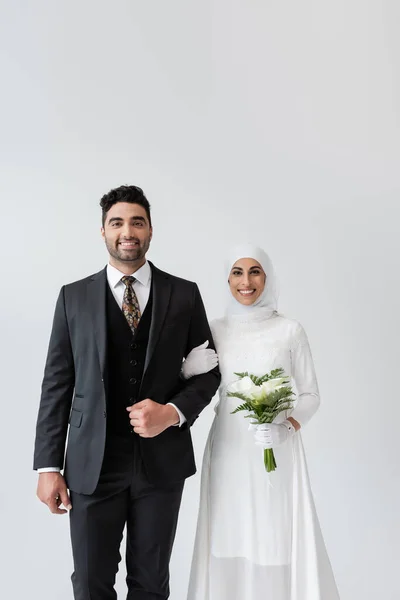 Happy muslim bride with bouquet standing near cheerful groom isolated on grey — Stock Photo