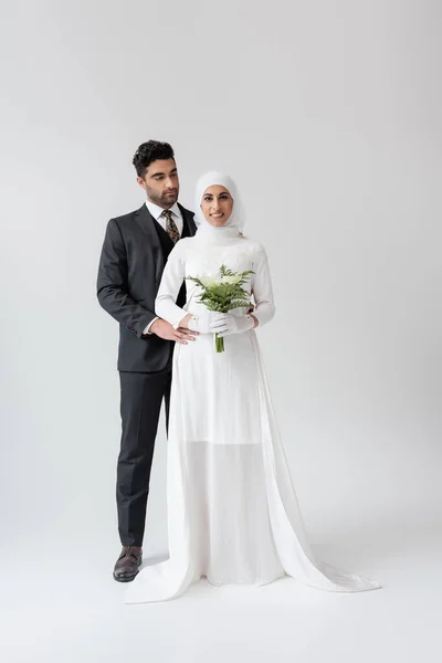 Marié musulman étreignant mariée heureuse en robe de mariée avec bouquet sur gris — Photo de stock