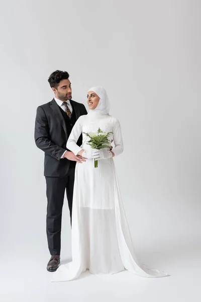 Marié musulman étreignant mariée gaie dans la robe de mariée avec bouquet sur gris — Photo de stock