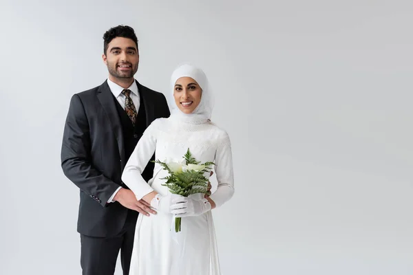 Мусульманский жених обнимает веселую невесту в свадебном платье с букетом, изолированным на сером — стоковое фото