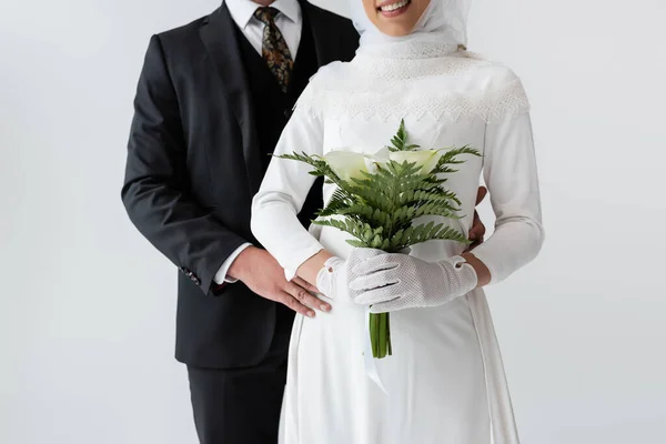 Cropped view of groom hugging happy bride in wedding dress with bouquet isolated on grey — Stock Photo