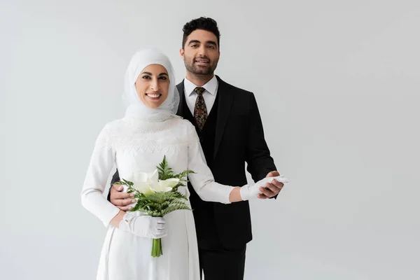 Sposo musulmano che tiene la mano della sposa felice in abito da sposa con bouquet isolato su grigio — Foto stock