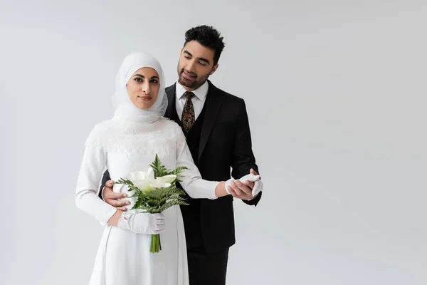 Мусульманский жених держит за руку улыбающуюся невесту в свадебном платье с букетом, изолированным на сером — стоковое фото