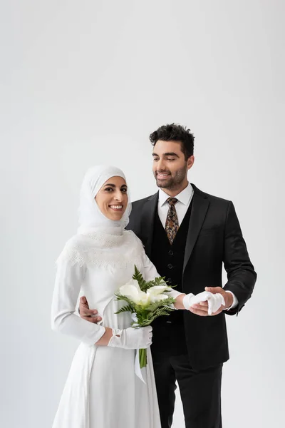Muslim groom holding hand of joyful bride in white dress with bouquet isolated on grey — Stock Photo