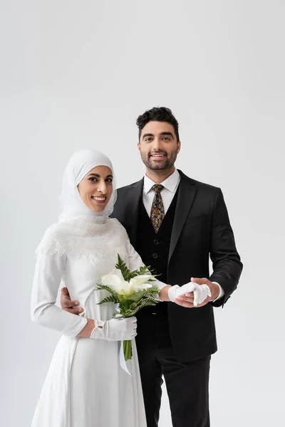 Muslimischer Bräutigam hält Hand einer glücklichen Braut im weißen Kleid mit Strauß isoliert auf grau — Stockfoto