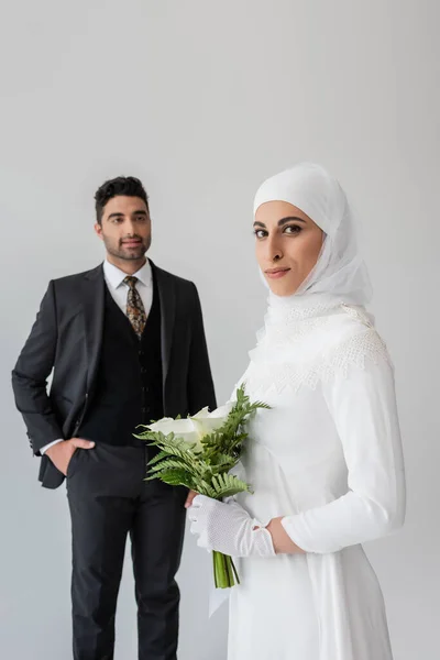 Homme musulman flou regardant jolie mariée avec des fleurs au premier plan isolé sur gris — Photo de stock