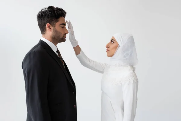 Vue latérale de la jeune mariée en hijab touchant le front du marié lors de la cérémonie de mariage isolé sur gris — Photo de stock