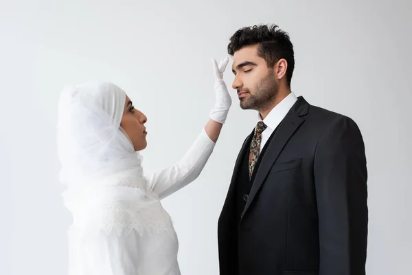 Young bride in hijab touching forehead of groom during wedding ceremony isolated on grey — Stock Photo