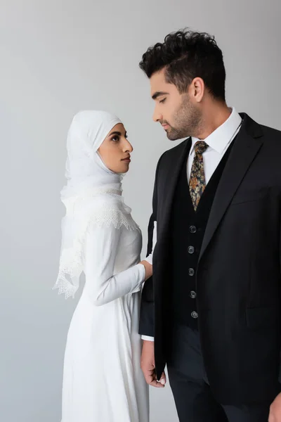 Donna in abito da sposa e hijab in piedi vicino al marito in abito isolato su grigio — Foto stock