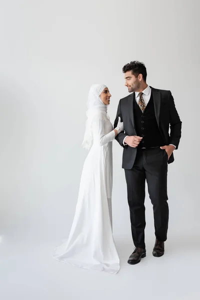 Sposo musulmano in piedi con mano in tasca vicino sposa felice in abito da sposa su grigio — Foto stock