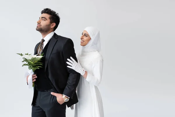 Glückliche muslimische Braut im Hijab umarmt Bräutigam mit Strauß isoliert auf grau — Stockfoto