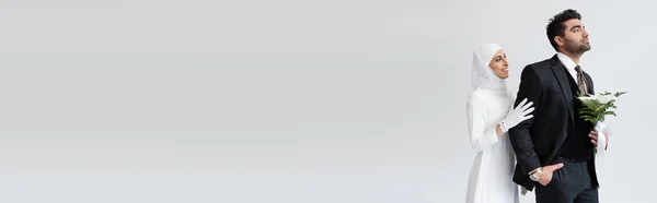 Щаслива мусульманська наречена в хіджабі обіймає нареченого з букетом ізольовано на сірому, банер — стокове фото