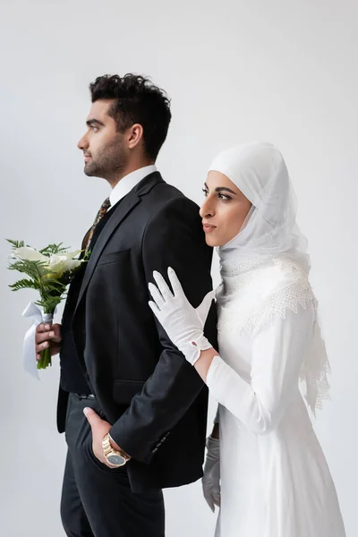 Muslim bride in hijab hugging groom with bouquet isolated on grey — Stock Photo