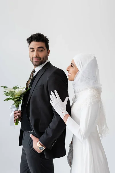 Happy muslim bride in hijab hugging groom with bouquet of calla lily flowers isolated on grey — Stock Photo