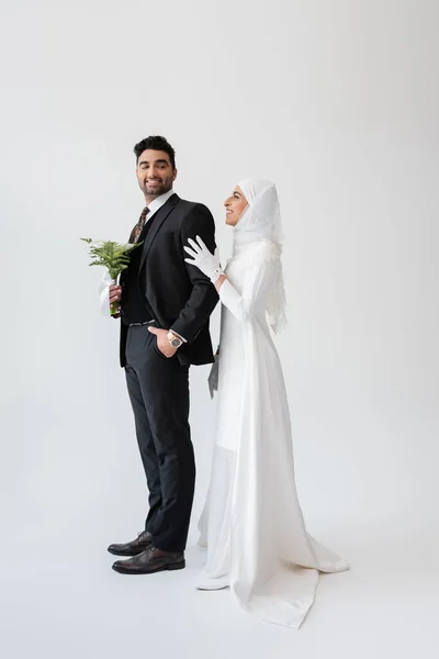 Счастливая мусульманская невеста в хиджабе обнимает жениха с букетом на сером — стоковое фото